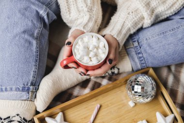 Genç bir kadın, rahat, yün, beyaz bir kazakla ekose üzerinde oturur ve elinde lokum dolu bir fincan kakao tutar. Hygge Yeni Yıl, tatil için samimi hazırlık. Mumlar, Noel topları ahşap tepside.