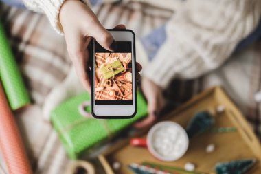 Akıllı telefonlu bir kadın sosyal ağlar için yılbaşı resmi ya da Noel resmi çekiyor. İçerik yaratılıyor. Tahta tepside kakao kupası şekerlemeler, oyuncak ağaç, mum, yıldızlar, paketlenmiş hediye..