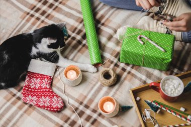 Genç kadın rahat örülmüş yün beyaz kazak, çorap ve Noel hediyesini puantiyeli ambalaj kağıdına sarar. Komik siyah beyaz smokinli kedi yakınlarda oynuyor..