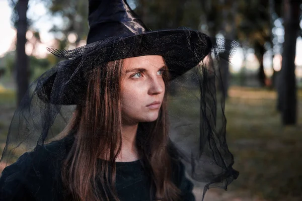 Jolie jeune belle femme en robe sombre et chapeau de sorcière debout au milieu des bois d'automne ou parc. Costume de fête d'Halloween. Portrait en gros plan. Ombre sur son visage. — Photo