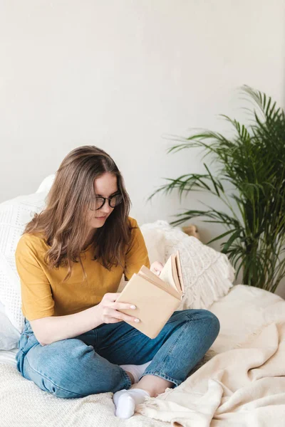 Ung vacker flicka student i glasögon, en orange T-shirt och blå jeans sitter på sängen med kuddar och läser pappersbok. Självskolekoncept hemma under karantänen. Hemundervisning, hobby. — Stockfoto