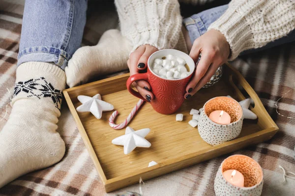 Ung kvinna sitter på rutig i mysig stickad ylletröja och håller i kopp kakao med marshmallows. Hygge nyår, mysig jul, förberedelser för semester. Trä bricka häxa ljus, stjärnor, bollar. — Stockfoto