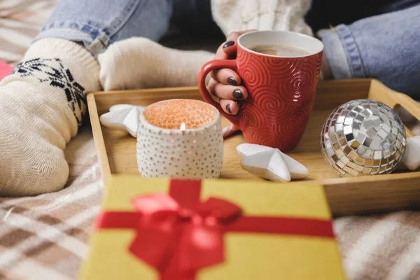 Young woman sits on bed in cozy knitted wool white sweater and holding gift-box with red gold. Hygge, New Years, Christmas, holiday preparations. Candles, Christmas balls, gifts, cocoa marshmallows.