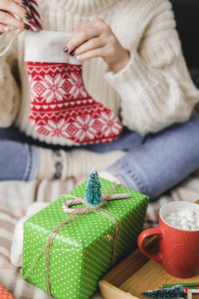 Young woman sits on plaid in cozy knitted woolen sweater and puts striped lollipop cane in Christmas sock for gifts. Preparing for winter holidays. Wooden tray with a mug of cocoa with marshmallows.