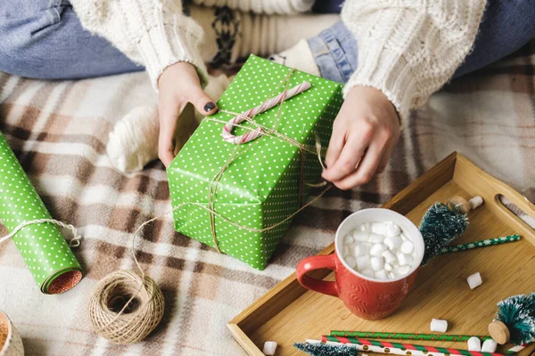 Молода жінка сидить на плетеному в'язаному вовняному білому светрі, шкарпетках і обгортає різдвяний подарунок у папері для обгортання крапок. Дерев'яний піднос з чашкою какао з зефіром, іграшковим деревом, свічкою, соломою . — стокове фото