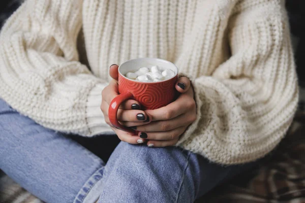 La jeune femme est assise sur le plaid dans un confortable pull blanc en laine tricoté et tient une tasse de cacao avec des guimauves dans ses mains. Hygge Nouvel An, Noël confortable, préparation pour les vacances. — Photo