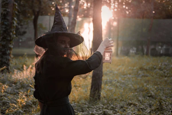 Jeune femme en robe sombre et chapeau de sorcières tient lanterne avec des bougies dans ses mains et éclaire la forêt. Costume de fête d'Halloween. Parc avec arbres d'automne. — Photo