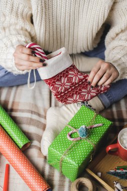 Genç kadın rahat yün bir kazakla ekose üzerinde oturuyor ve hediye olarak Noel çorabının içine çizgili lolipop kamışı koyuyor. Kış tatiline hazırlanıyorum. Marşmelovlu bir kupa kakaolu ahşap tepsi..