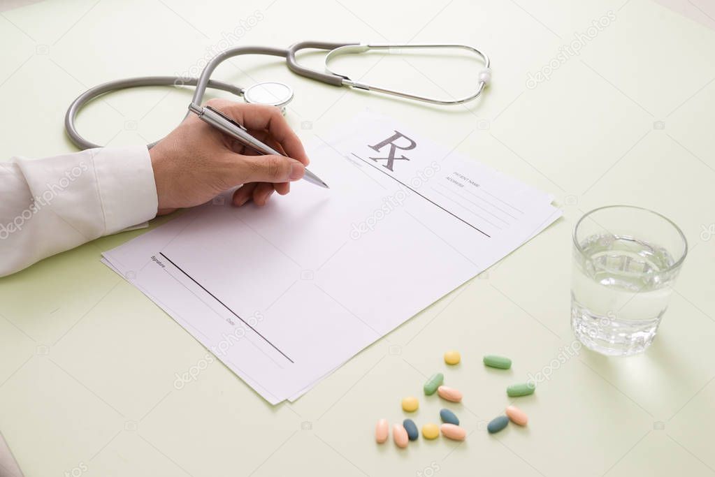 Top view of male doctor hand writing prescription over table with pills, glass of water and stethoscope 