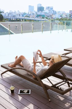 Genç Asyalı adam bir güneşli yaz gününde havuzun yanında kitap okuma