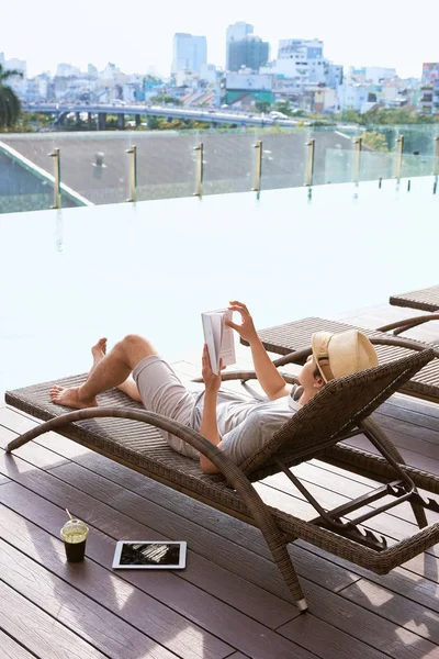 Genç Asyalı adam bir güneşli yaz gününde havuzun yanında kitap okuma