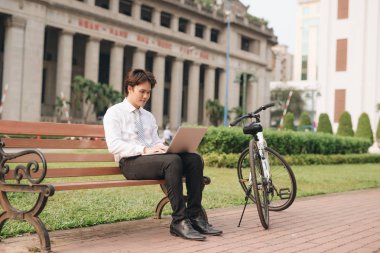 Dizüstü bilgisayarı ve bisikleti olan yakışıklı genç işadamı parkta bankta oturuyor.