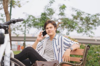 Bisiklet sürdükten sonra parkta bankta oturan genç yakışıklı işadamı cep telefonuyla konuşuyor.