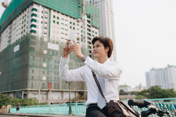 Young man taking a photo with his smart phone while standing on the city street with his bicycle beside him