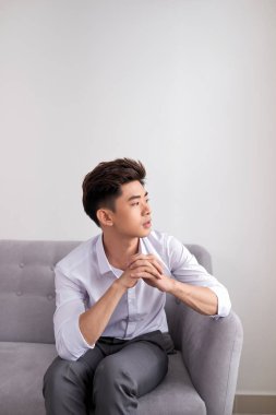Oturan genç Asyalı adam kanepe ve düşünce bir şey, üzerinde beyaz izole görünür