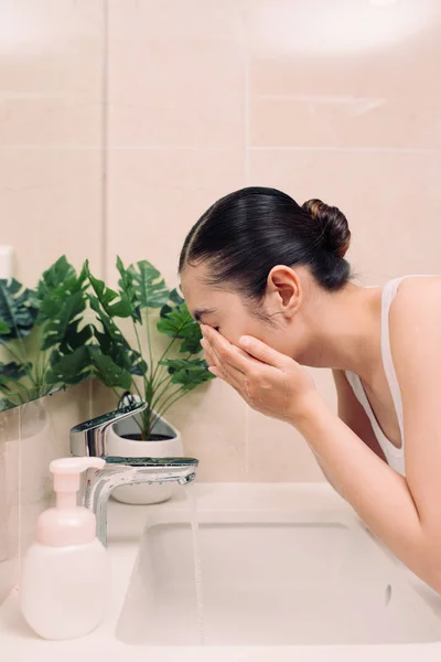 Woman washing her face with water above bathroom sink.