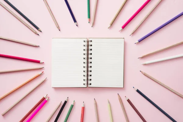 笔记本在中心与明亮的彩色铅笔衬里周围的粉红色背景 — 图库照片