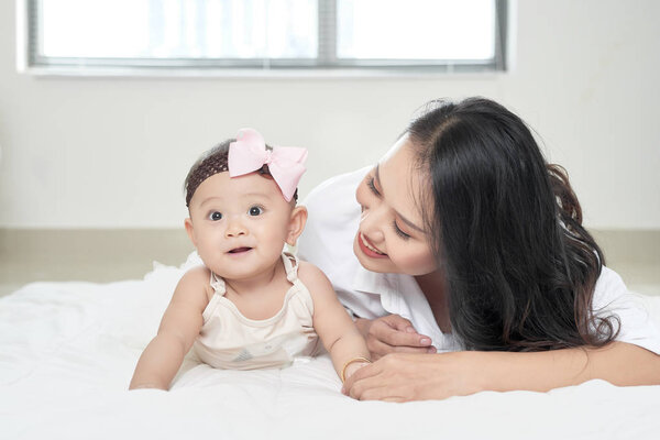 Mother and baby daughter happy and beautiful home together playing on bed on window background