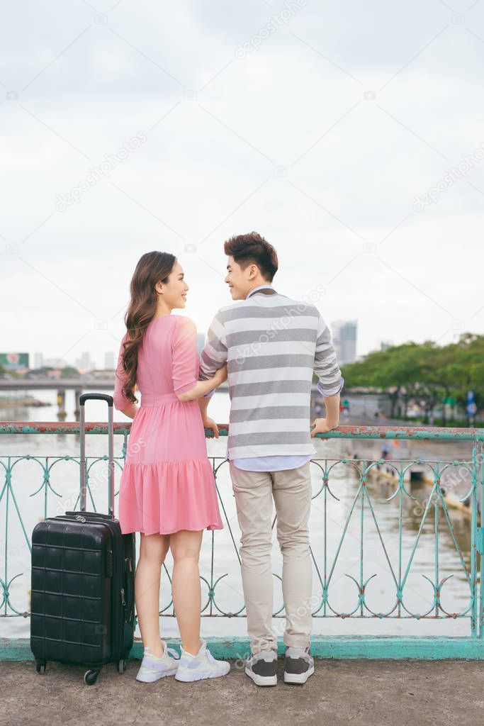 back view of young asian lovers with suitcase standing on bridge over river with cityscape on background, travel concept  
