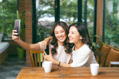 Two young attractive women taking selfie with smartphone while sitting in cafe clipart