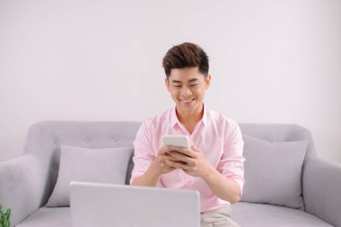 Kanepe dizüstü bilgisayar ile mesaj ile smartphone üzerinde oturan genç gülümseyen Asyalı adam