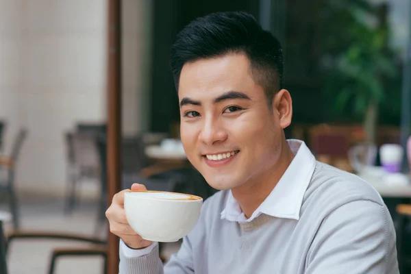 Young Asian man holding coffee cup and looking at camera while sitting in cafe