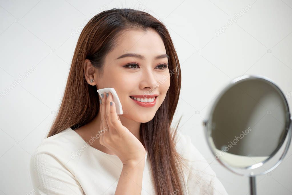 Beautiful cheerful young woman using cotton disk and looking at reflection in mirror with at dressing table