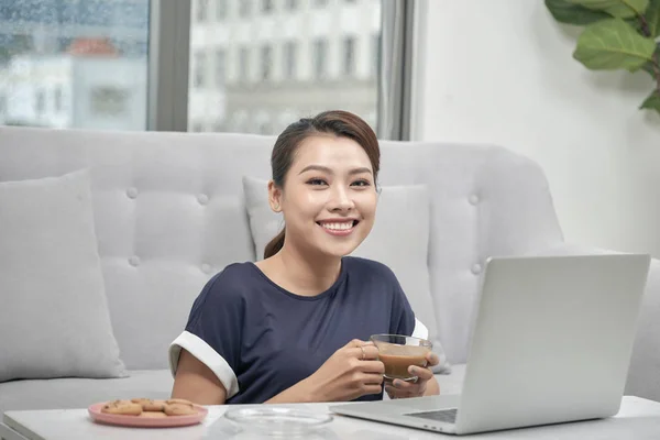 ラップトップ コンピューターを自宅で座って一杯のコーヒーを保持している笑顔の若いアジア女性 — ストック写真