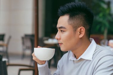 Yeni gün kafede başlangıç. Kahve fincanı tutarak ve uzak kafede otururken arıyorum Genç Asyalı adam yan görünüm