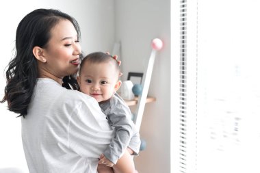 Güzel genç anne şirin bebeği ile oynama ve evde pencere yanında dururken gülümseyen