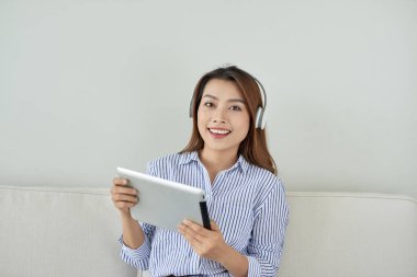 Çok gülümseyen genç kadın kanepe ve usinging tablet üzerinde oturan kulaklıklar giymek.