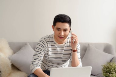 Asya genç adam evde kanepede smartphone ve dizüstü bilgisayar ile çalışma.