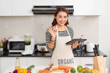 Güzel ve genç birbayan dijital tablet kullanma ve evde mutfakta yemek yaparken gülümseyen