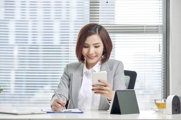 Businesswoman making notes looking at a cellphone at office. Woman entrepreneur sitting at the table writing notes while working on cellphone.