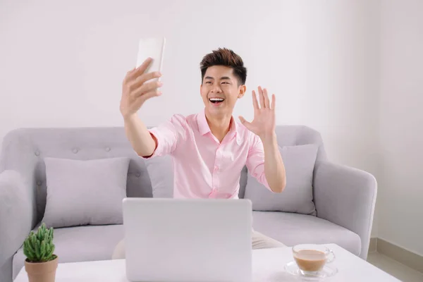 Young handsome asian man with laptop, taking a selfie with phone sitting on a couch at home