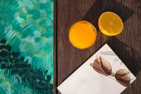 Orange fresh juice, book and sunglasses near pool