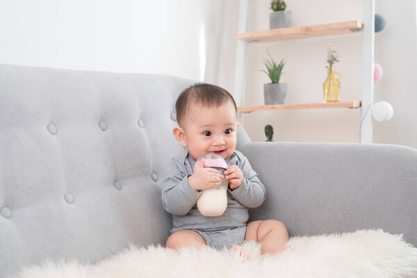 Little cute baby girl sitting in room on sofa drinking milk from bottle and smiling. Happy infant. Family people indoor Interior concepts. Childhood best time!