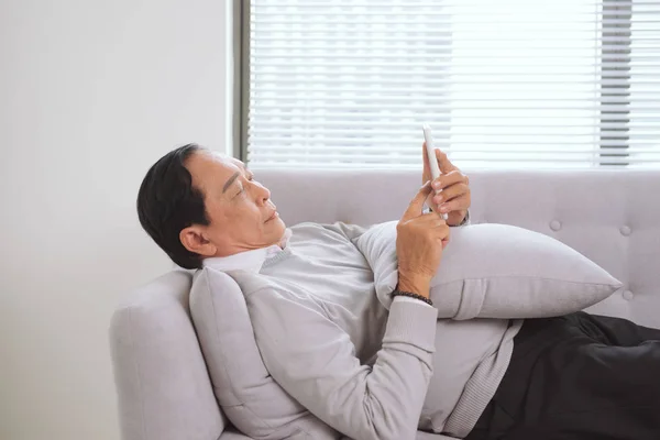 Kıdemli Asya Adam Kanepede Yatıyor Telefonda Bakıyor — Stok fotoğraf