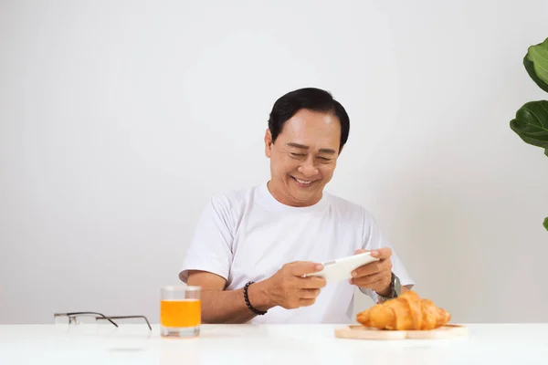 快乐的资深夫妇在家里有羊角面包早餐和喝橙汁 使用智能手机 — 图库照片