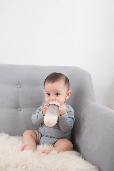 Little cute baby girl sitting in room on sofa drinking milk from bottle and smiling. Happy infant. Family people indoor Interior concepts. Childhood best time!