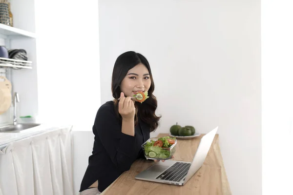 Young woman with salad working on laptop in the kitchen