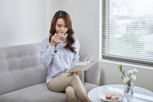 Young attractive woman read book and drink coffee sitting near window at home