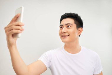 Gülümseyen genç Asyalı adam beyaz arka plan üzerinde akıllı telefonda selfie fotoğrafı çekiyor