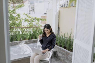 Sabah kahvesini zevk. Çay bardağı tutan ve onun açık evin terasında bir sandalyede otururken kameraya bakarak güzel genç gülümseyen kadın