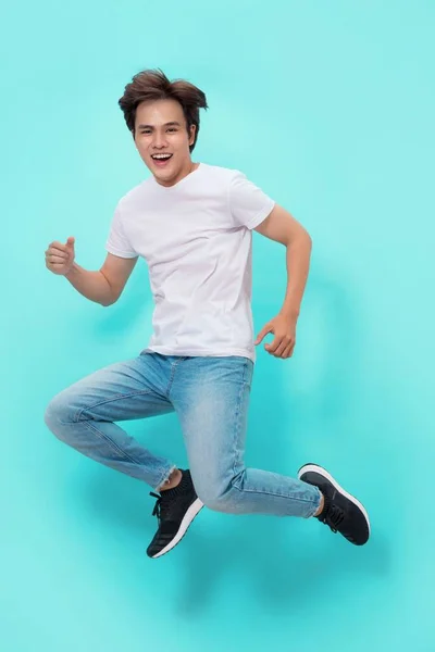 Full length portrait of an excited young man in white t-shirt jumping while celebrating success isolated over blue background