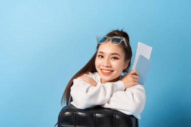 Genç Asya kadın oturma yakın bir bavul Holding pasaport üzerinde bir mavi arka plan.