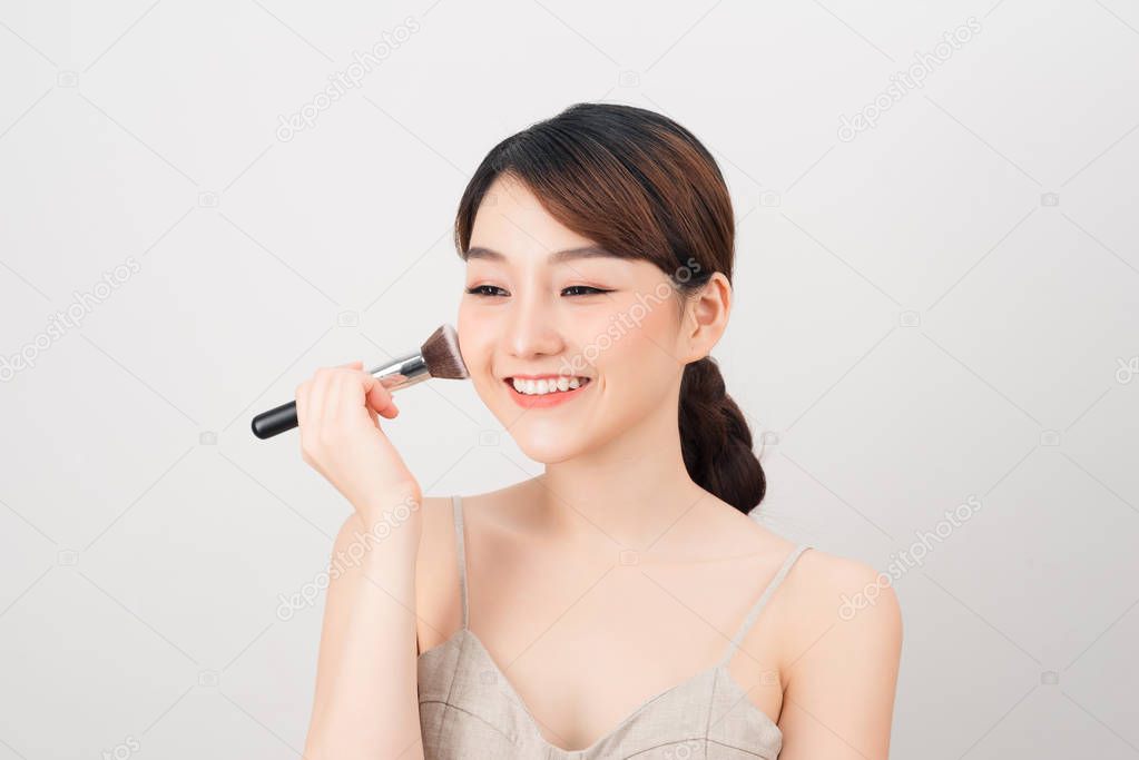 Cheerful woman is doing make up on her face using brush. Beauty routine on white background.