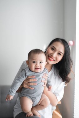 Güzel genç anne şirin bebeği ile oynama ve evde pencere yanında dururken gülümseyen