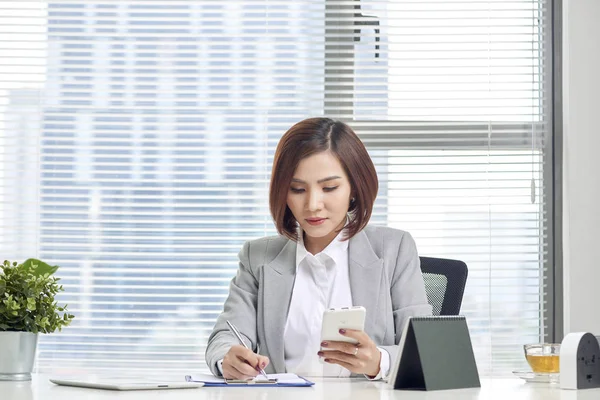 Businesswoman making notes looking at a cellphone at office. Woman entrepreneur sitting at the table writing notes while working on cellphone.