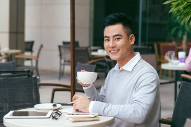 Sabahının tadını çıkarıyor. Kahve fincanı tutan ve kafede otururken gülümseyen kameraya bakan asyalı genç adam - Resim
